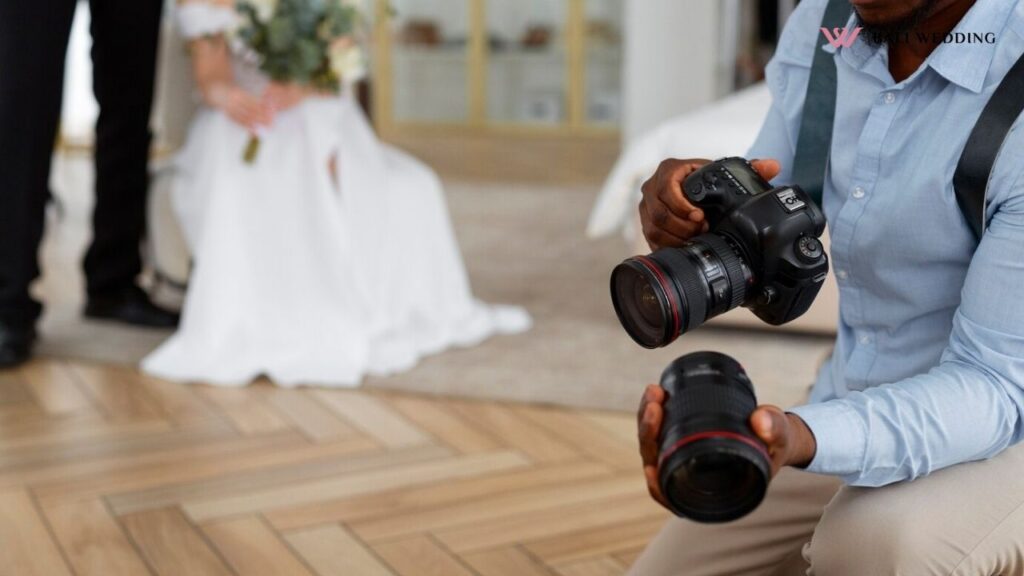 A photographer kneeling down, holding two professional cameras in preparation for a shot. In the blurred background, a bride in a white gown sits holding her bouquet, while another figure stands nearby.