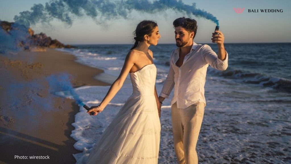 A bride and groom standing on the beach, holding blue smoke flares as they gaze into each other's eyes. The scene features waves gently crashing on the shore, creating a romantic, sunset-lit backdrop.