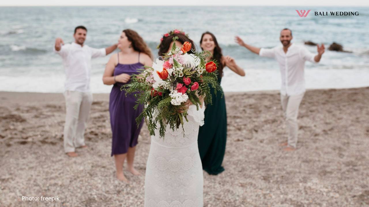 Bride with a large bouquet of colorful flowers standing on a beach with friends celebrating in the background.