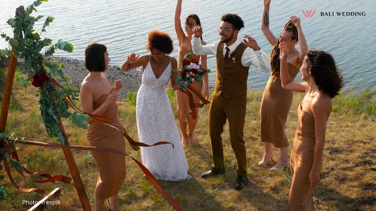 Bride and groom dancing joyfully with bridesmaids by a scenic lake at an outdoor wedding ceremony.